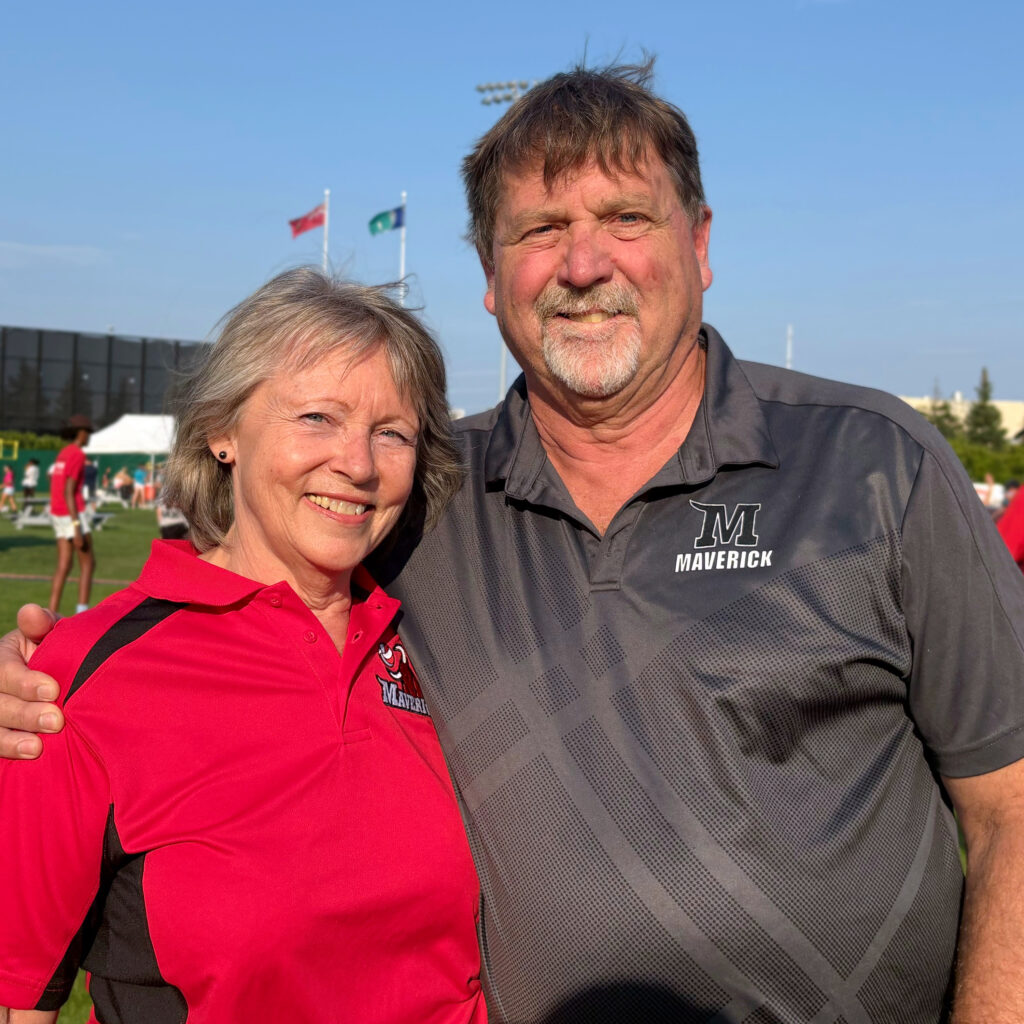 Kerry and Chris MacLean pose side by side at an outdoor sporting event. Kerry is wearing a grey Mavericks polo shirt.