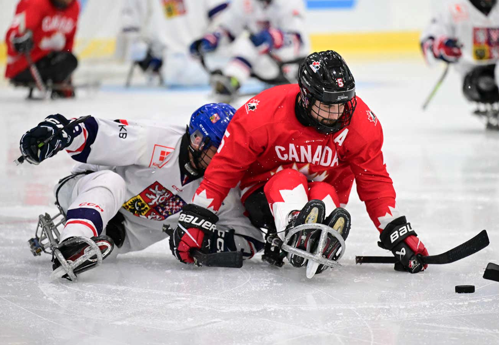 Tyrone Henry reaches to play the puck which sits in front of his left stick, while he holds off a player from Czechia on his right side. Henry wears the A on his red Canada jersey.