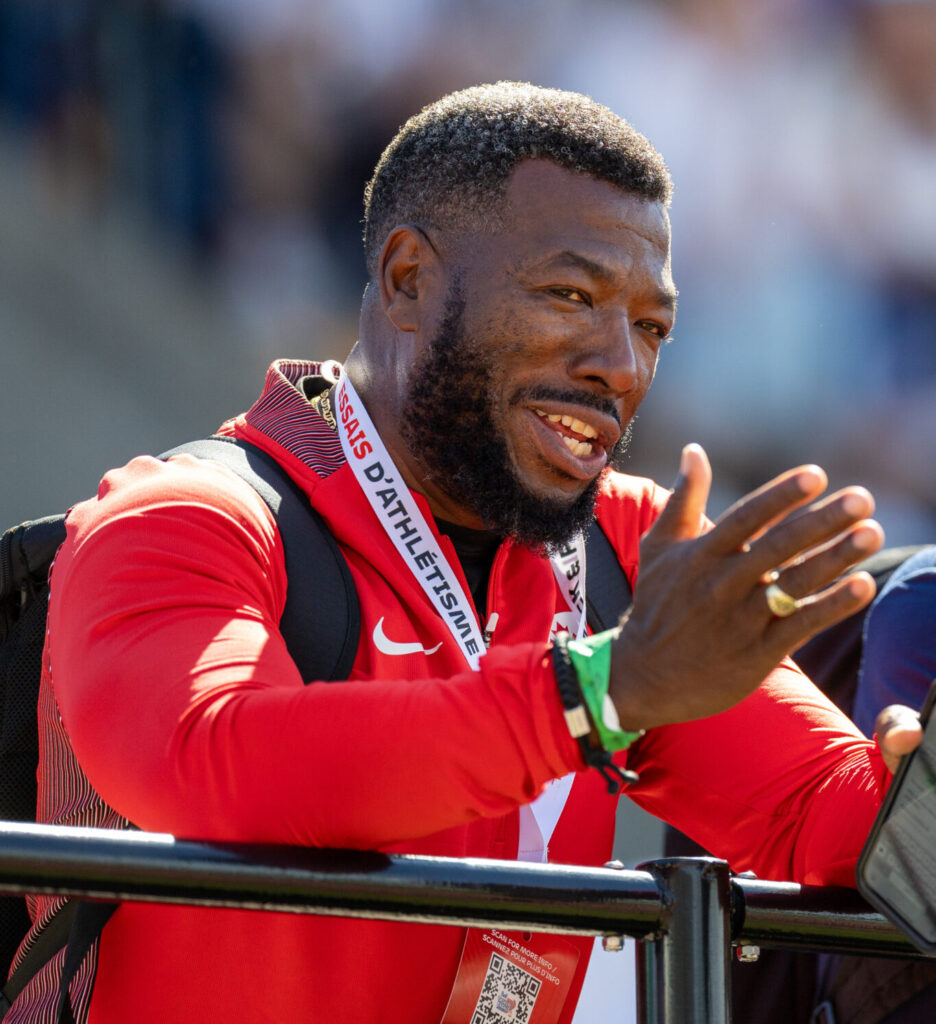 Glenroy Gilbert gestures with his right hand while chatting with a fellow coach along the sidelines of a track and field event.